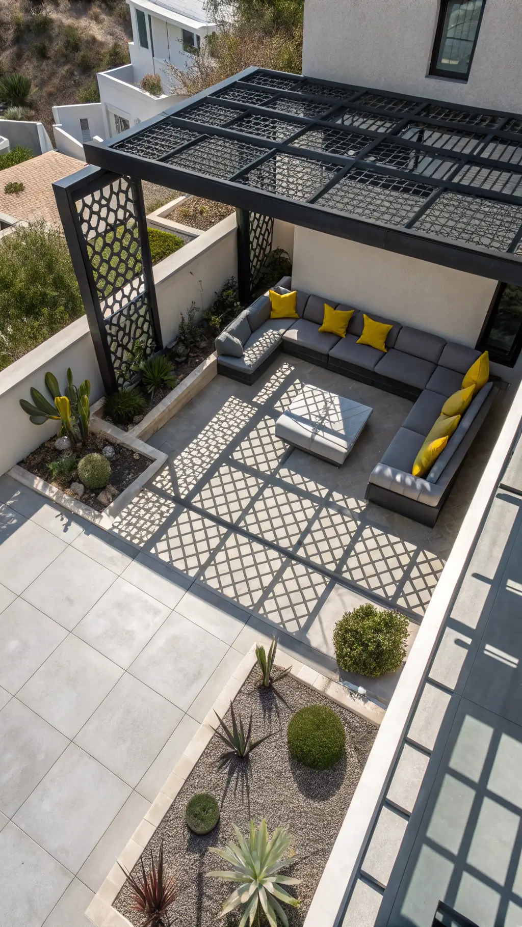 Overhead drone view of a modern patio with geometric lattice panel cover, contemporary gray sectional seating with yellow pillows, and stainless steel planters with succulents under midday light.