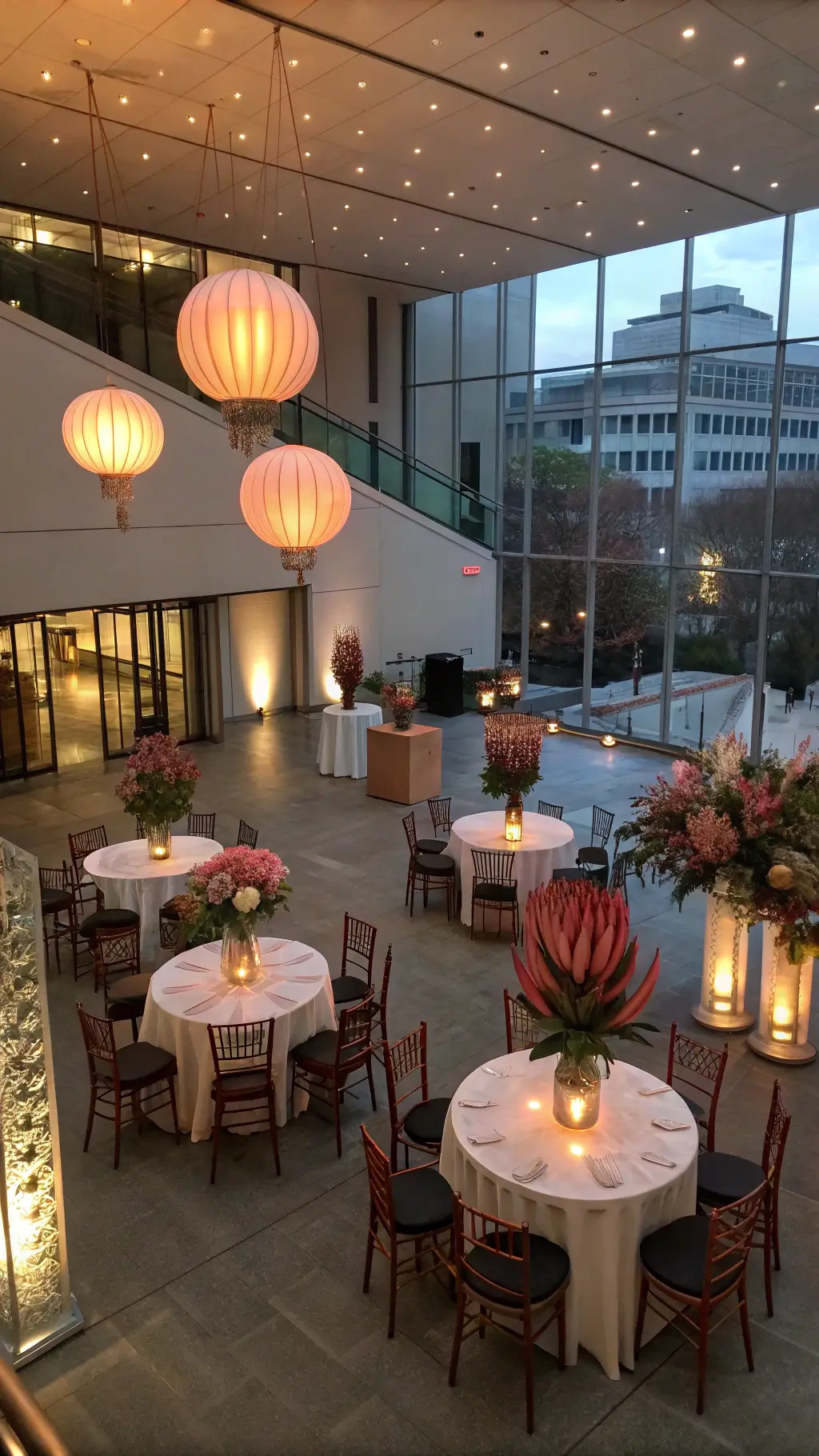 Modern art installations as dynamic backdrop in a contemporary gallery space, adorned with high cocktail tables in champagne linens, copper chairs, and geometric floral arrangements featuring protea and anthuriums, all illuminated by a warm glow from floating paper lanterns at dusk.